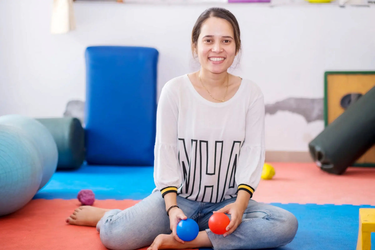Smiling woman holding therapy balls, seated in a brightly colored fitness studio.
