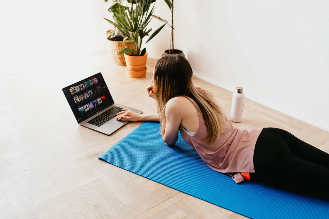 From above side view of young long haired female in casual wear lying on floor on blue mat while using laptop at home