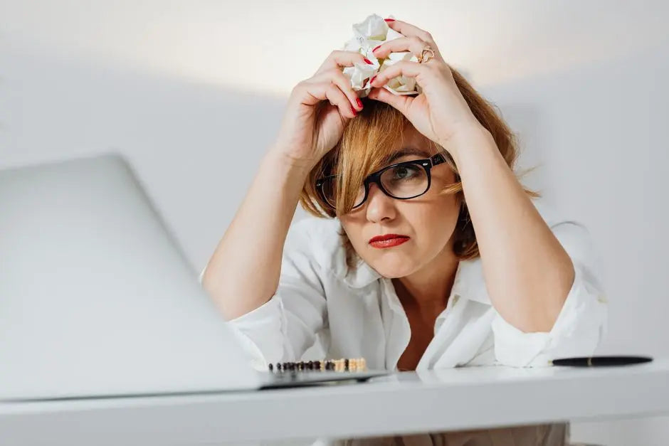 Frustrated woman with eyeglasses at a desk, crumpling paper while using a laptop indoors.