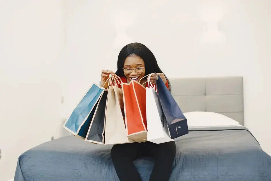 Happy woman holding colorful shopping bags, seated on a bed in a cozy bedroom.