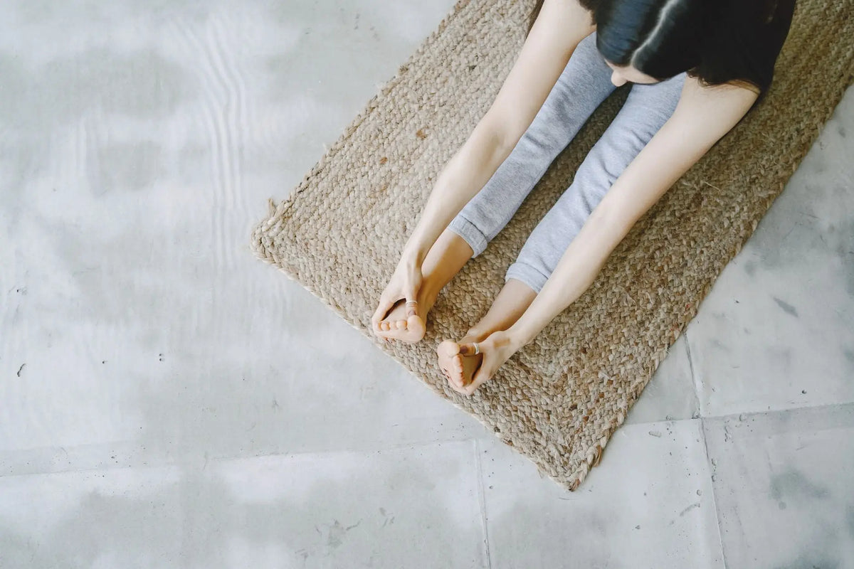 Top view of a woman practicing seated forward bend on a woven mat indoors.
