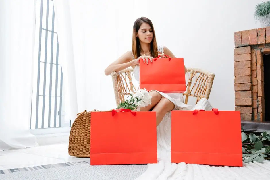 Woman sitting in a stylish chair with red shopping bags and flowers in a cozy indoor setting.