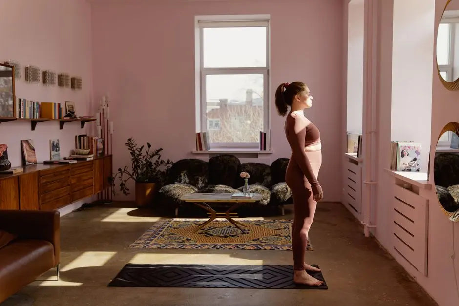 A woman stands on a yoga mat indoors, bathed in natural light from a window, practicing yoga.