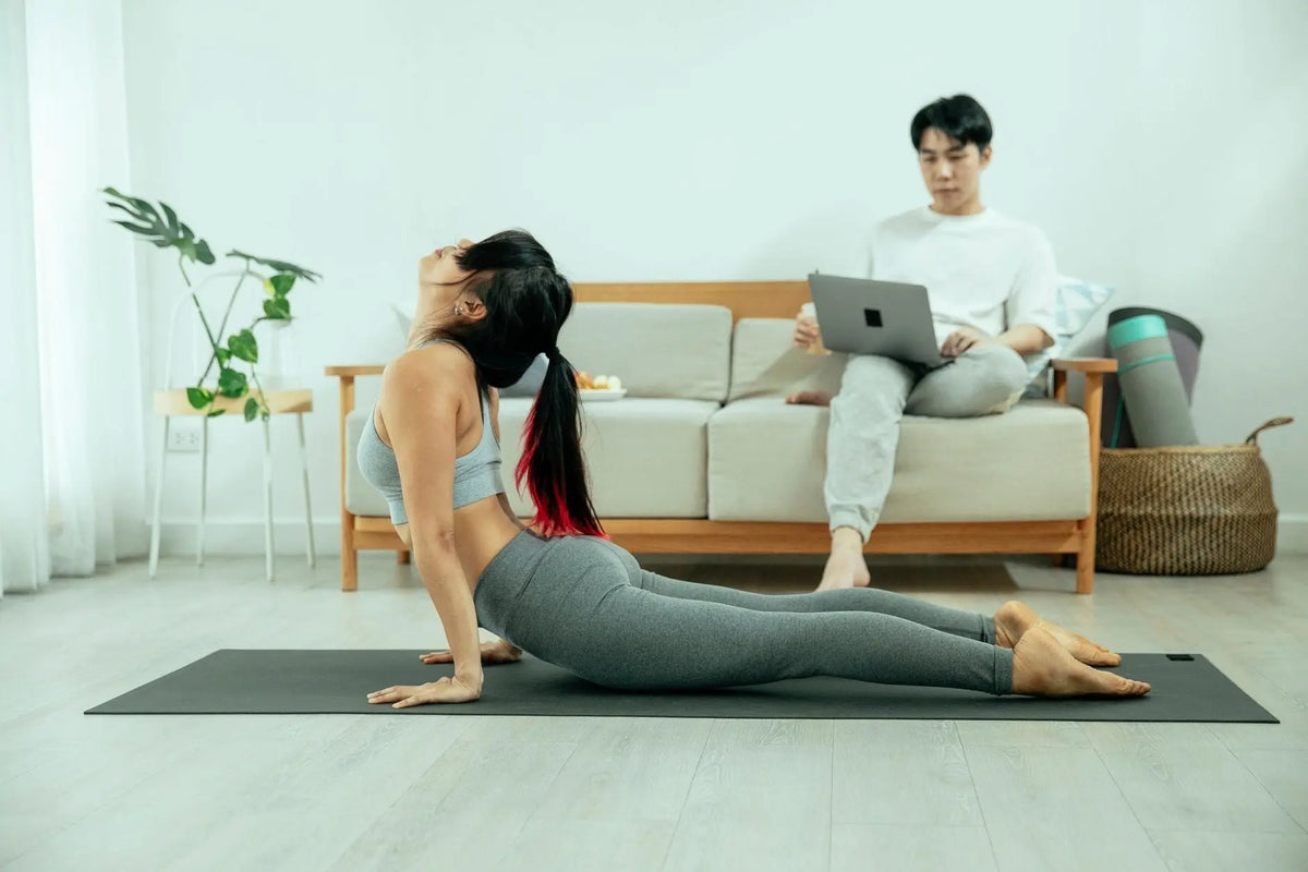 Full body of female in sportswear stretching body on mat while Asian male sitting on couch and using laptop at home