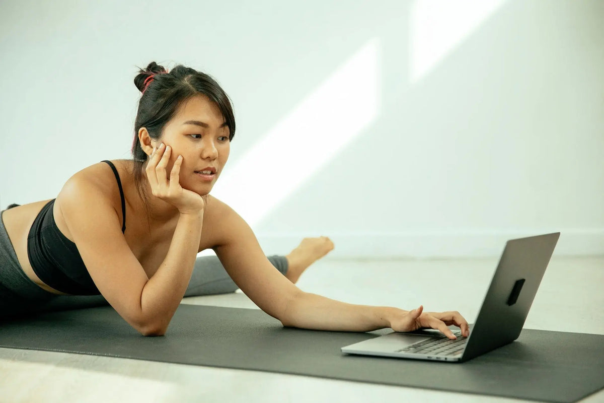 Charming Asian female in sportswear stretching legs while using laptop on mat in light apartment