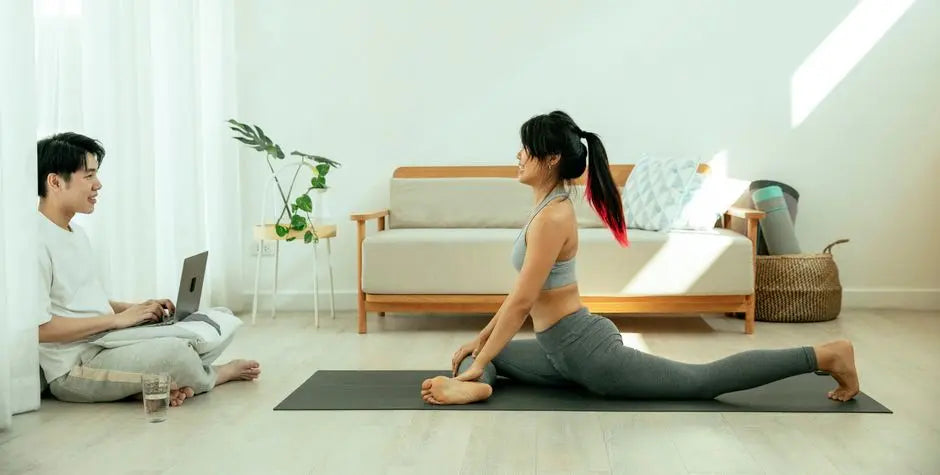 Side view of concentrated barefoot female in sportswear performing Half Pigeon pose on mat during yoga session and smiling male sitting on floor crossed legs with netbook