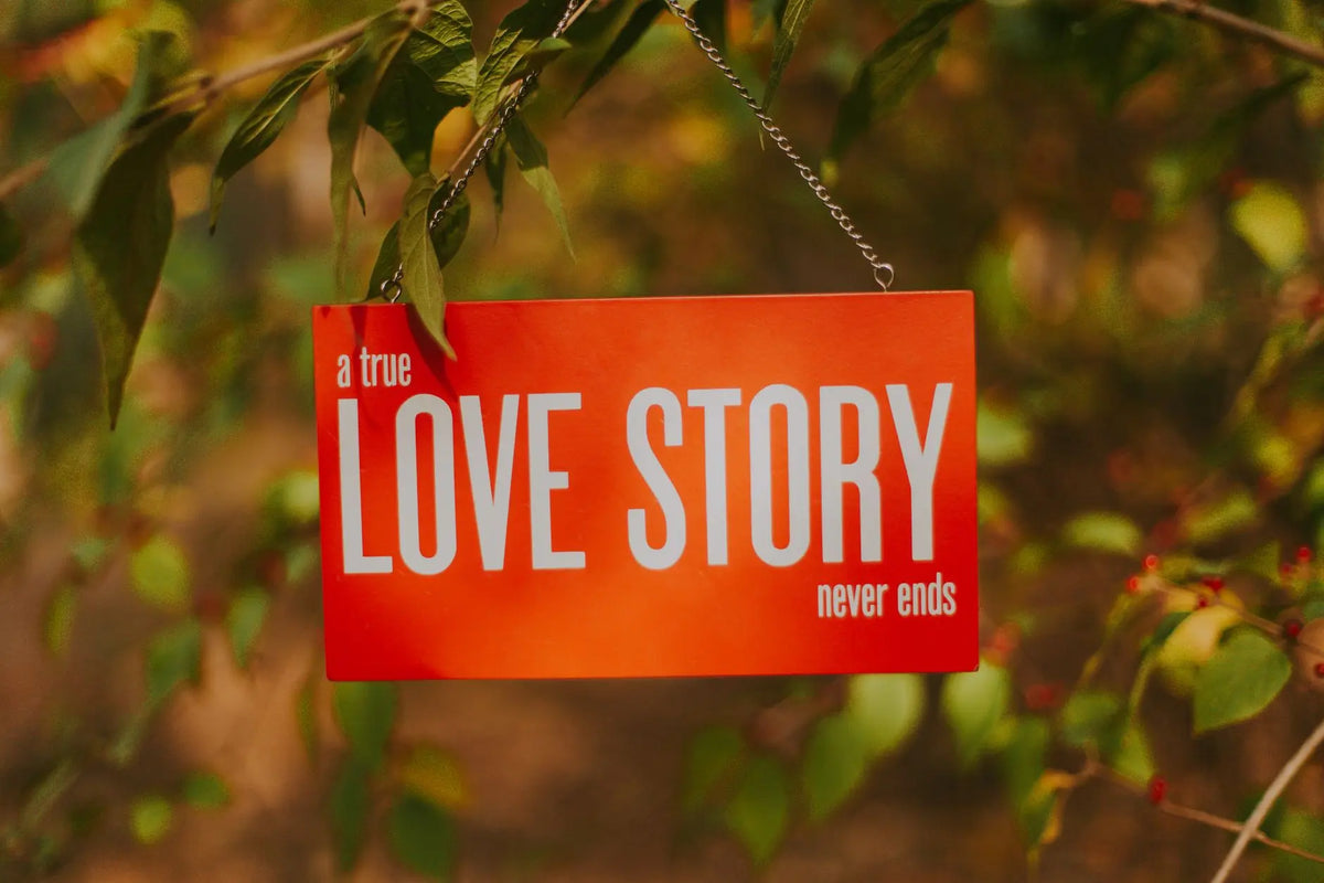 Orange sign stating 'A true love story never ends' hanging from a tree branch outdoors.